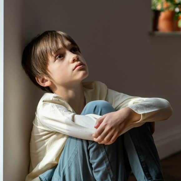 Lonely boy sadly looking up hugging knees sits near window at home alone. Kid psychological trauma