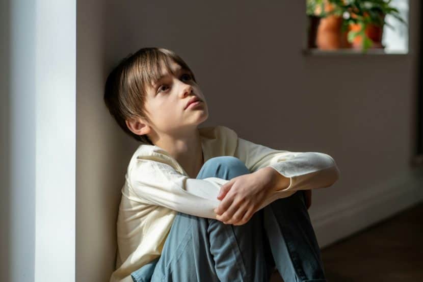 Childhood trauma and neglect can be passed on to future generations Lonely boy sadly looking up hugging knees sits near window at home alone. Kid psychological trauma